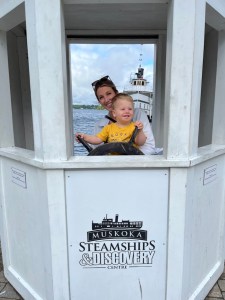 Daughter and grandson in a small replica of Segwun, grandson is on his moms arms and pretending to hold the wheel of the ship.
